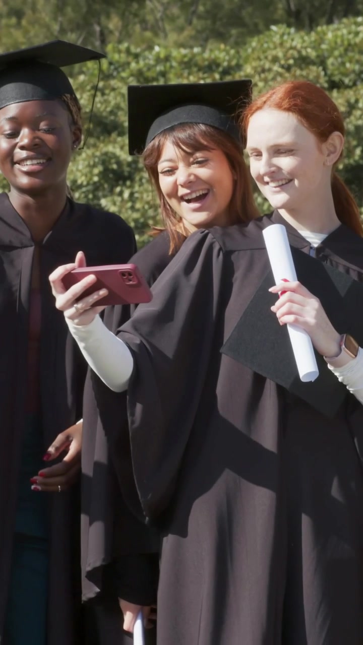 Video verticale: Un laureato con cappello alza il diploma ed esorta un gruppo a fare un selfie con un telefono rosa nel campus.
