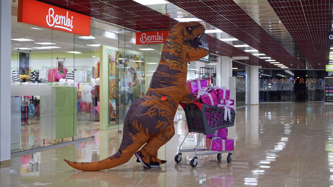 Orange T-Rex is pushing the shopping cart filled with presents. Man in dinosaur costume moving quickly by the hall of shopping mall.