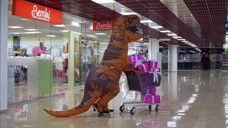 orange t-rex is pushing the shopping cart filled with presents. man in dinosaur costume moving quickly by the hall of shopping mall.