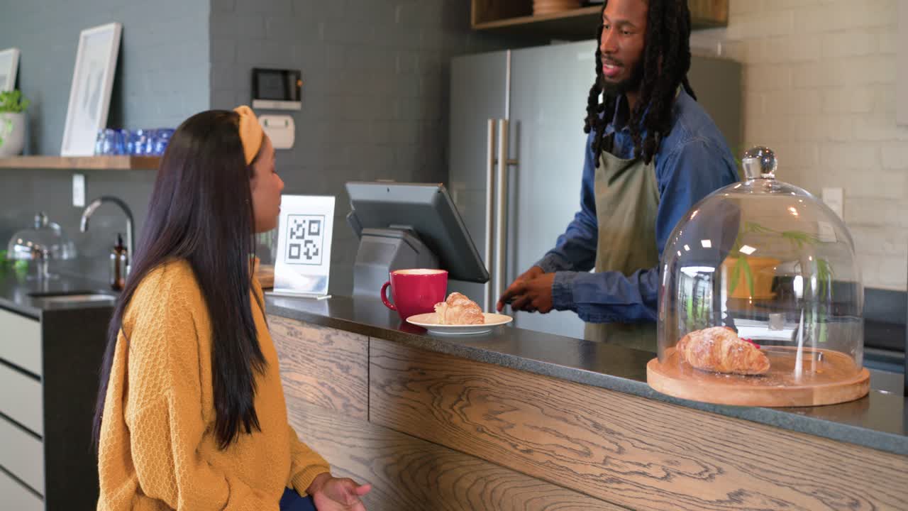 インド人女性がカフェに入り、バーでバリスタがクロワッサンとコーヒーを提供し、女性がそれらをすすりながら支払いを済ませる。