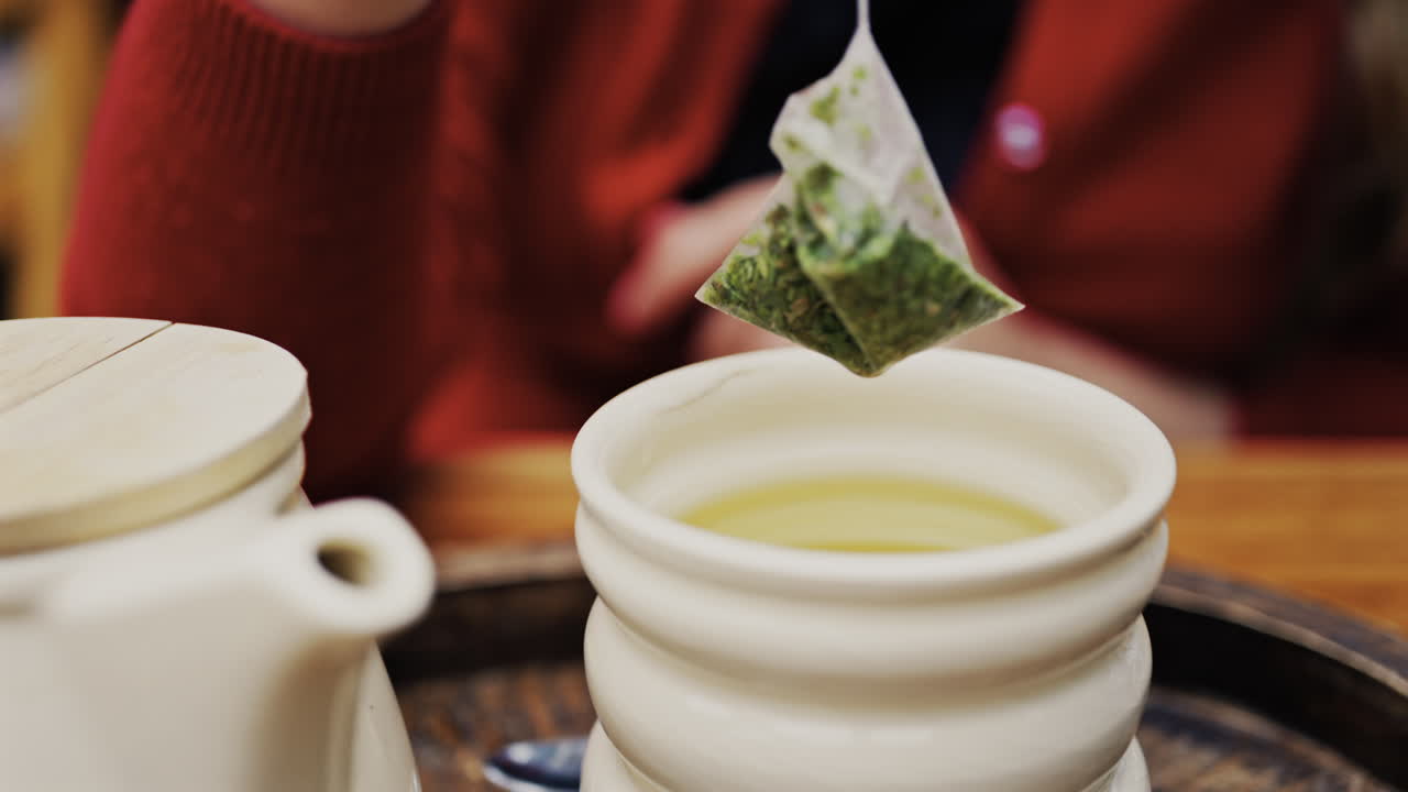 Close up of a woman in a red suit dipping a green tea bag in hot water in a white cup