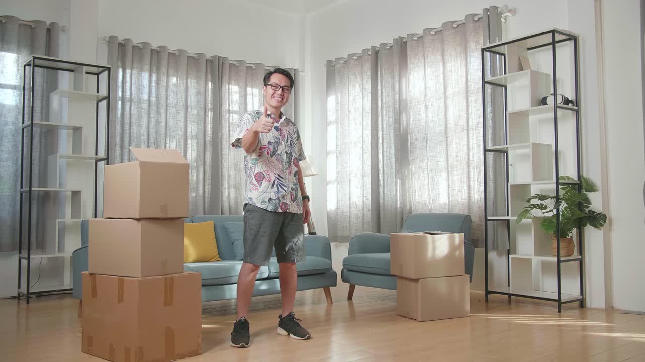 hombre asiático con cajas de cartón sonriendo y mostrando los pulgares hacia arriba gestos a la cámara en la nueva casa