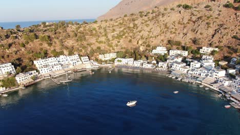 Drone-view-in-Greece-flying-over-blue-sea-in-Loutro-small-white-house-town-and-small-boats-next-to-a-hill-on-a-sunny-day