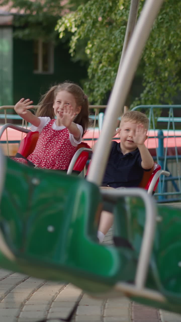 Happy little children wave hands riding carousel in theme park slow motion. Brother and elder sister have fun in amusement park. Urban safe entertainment