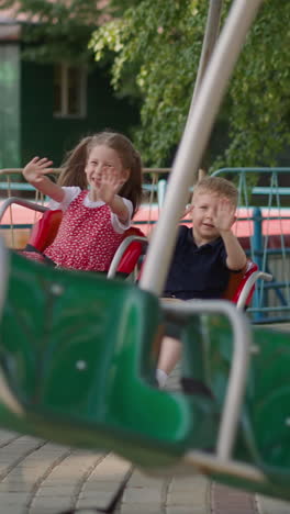 happy little children wave hands riding carousel in theme park slow motion. brother and elder sister have fun in amusement park. urban safe entertainment
