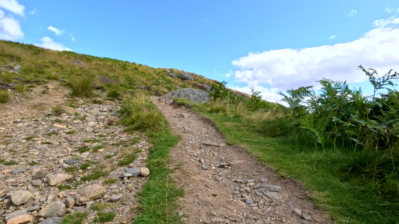 A steady walk up a rugged, rocky hillside trail bordered by grass and shrubs under bright daylight, with smooth handheld camera movement and wide landscape views