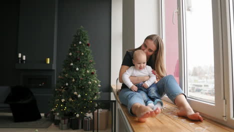 Young-mother-is-holding-her-son's-hands-while-he-is-walking-on-the-window-sill-decorated-with-Christmas-wreath.-Happy-family-kissing