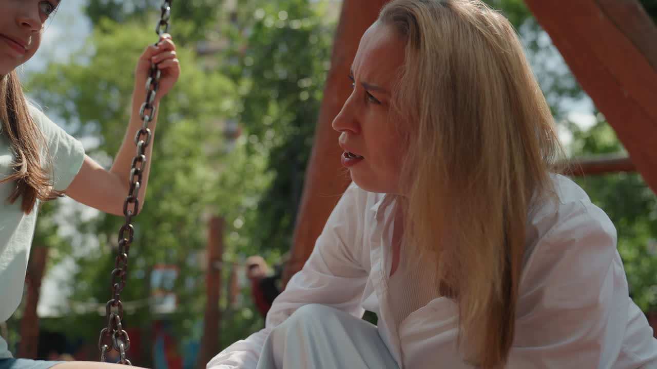 Vit mamma som undersöker dotterns ben på en gunga, intim parkscen med mjukt solljus, gungställning i trä, grön lövbakgrund, mamma knäböjer bredvid barnet, bekymrat uttryck, försiktiga händer.