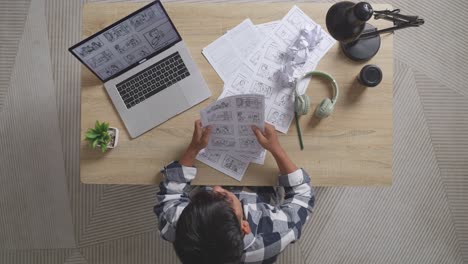 vista superior de un artista masculino asiático estresado mira su boceto luego lanza papeles mientras dibuja el storyboard en la mesa con una computadora portátil, auriculares y papeles arrugados en el estudio