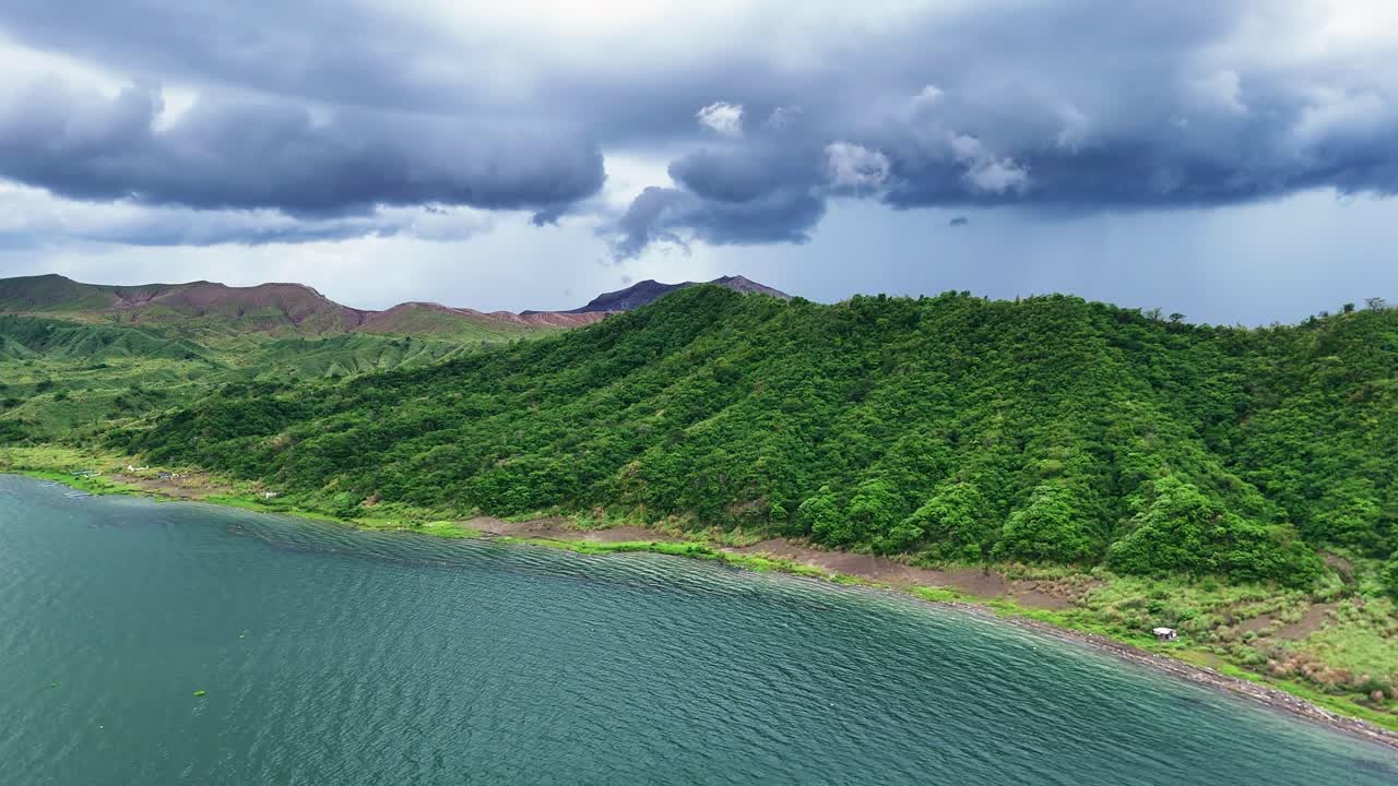 Aerial rise from Taal Lake, sweeping over the verdant slopes of the Tagaytay mountains, revealing Taal Volcano under a stormy sky, with dark clouds enhancing the dramatic scenery.