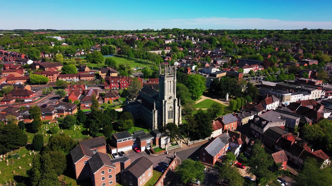 Amplias imágenes aéreas de dron de la Iglesia de St Mary en Andover, mostrando la histórica torre de la iglesia, el cementerio, los tejados circundantes, casas, carreteras, espacios verdes y el paisaje urbano más amplio de Hampshire bajo un cielo despejado.
