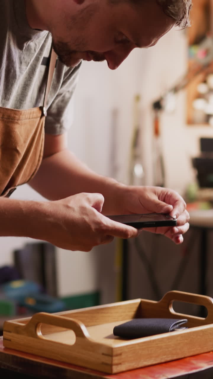 Bearded man takes photo of stylish leather purse with cellphone on tray in studio closeup. Master of manual manufacture presents product