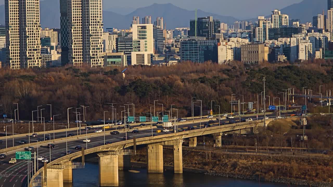 Traffico sulla rampa curva dell'autostrada Gangbyeonbuk-ro con lo skyline della città di Seul e le montagne sullo sfondo.