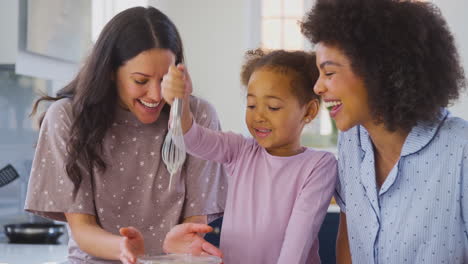 familia con dos madres vestidas con pijama haciendo panqueques matutinos en la cocina en casa con la hija