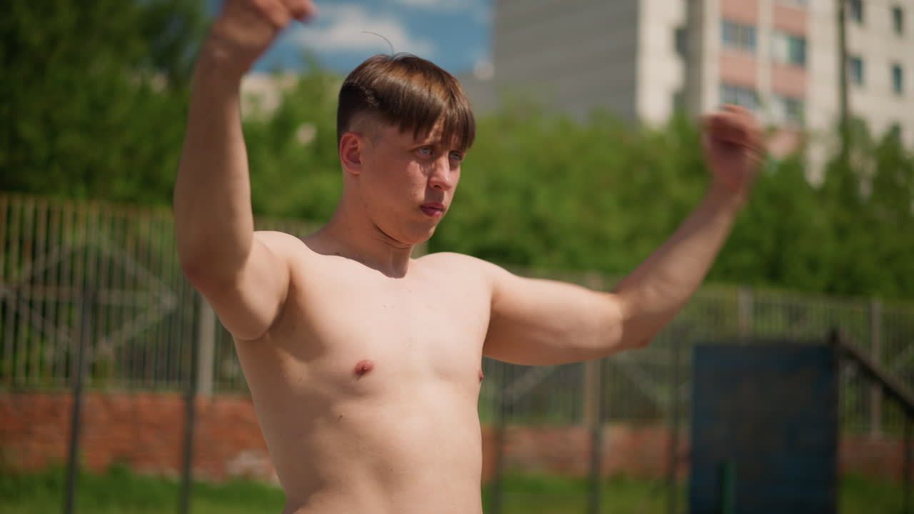 옷을 벗은 남자가 펀치를 연습하고, 흰색 피부의 남자가 도시의 녹색 배경 근처에서 펀치 기술을 연습하며, 셔츠를 입지 않은 남자가 아파트 건물과 무성한 나무 앞에서 펀치 훈련을 하고 있어요.