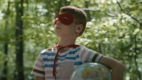 Ginger-caucasian-boy-with-superhero-costume-holding-globe-in-the-forest.