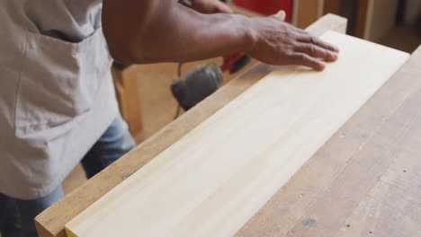 carpintero afroamericano que lleva una máscara protectora lijando una tabla de madera con una lijadora eléctrica
