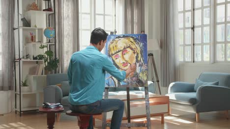 hind view of an asian artist man sitting on round wooden chair without backrest holding paintbrush mixed colour and painting a girl's face on the canvas