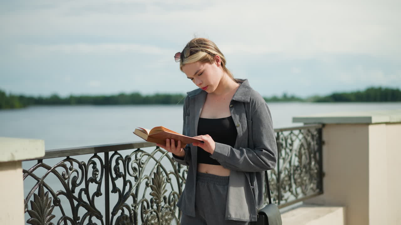 Young lady in grey clothing and black handbag standing close to a fence reading a book with focus as the wind blows the pages and her hair, trees are visible in the distance and a riverfront