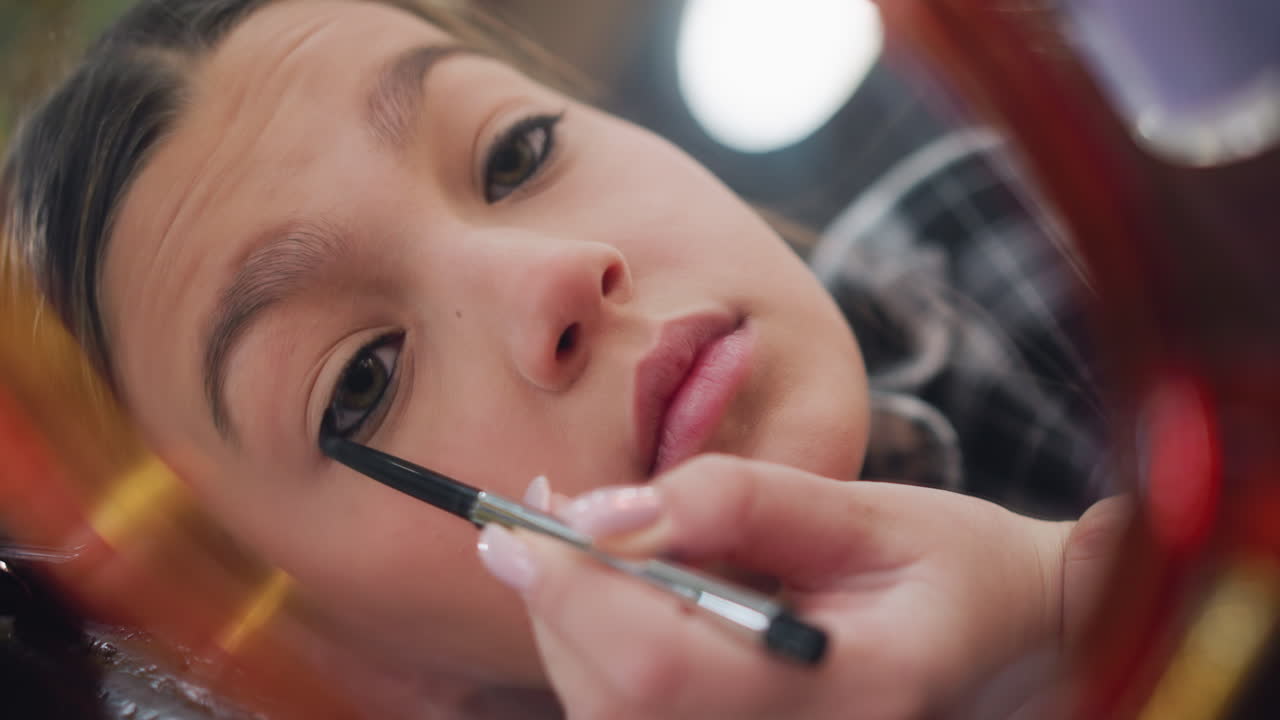 Mirror view of young lady carefully applying eye pencil to eyelid, capturing focus and precision, close-up on delicate makeup process highlighting attention to detail and grooming routine