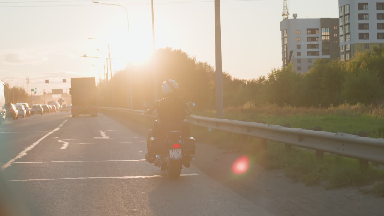 Motorcyclist with passenger rides along highway into glowing sunset with bright sunlight illuminating road, vehicles moving in distance and warm golden light