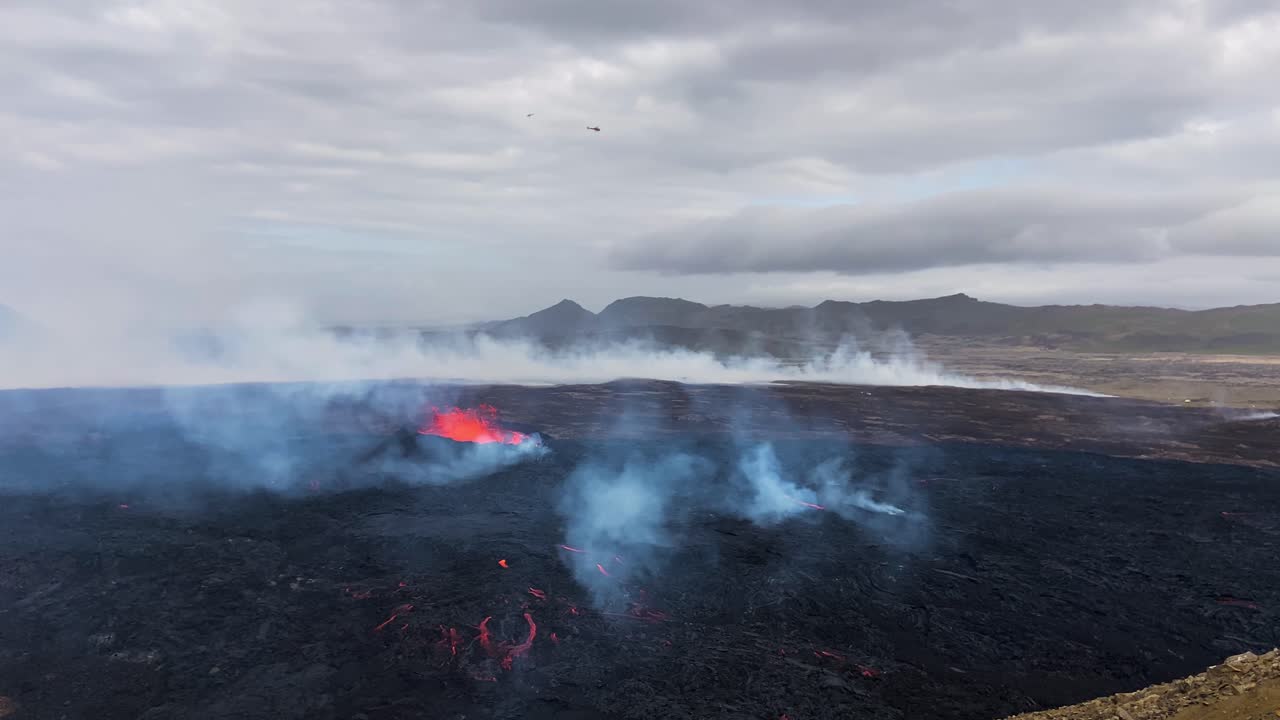 Lava flows and smoky plumes from Fagradalsfjall under a cloudy sky with a helicopter overhead