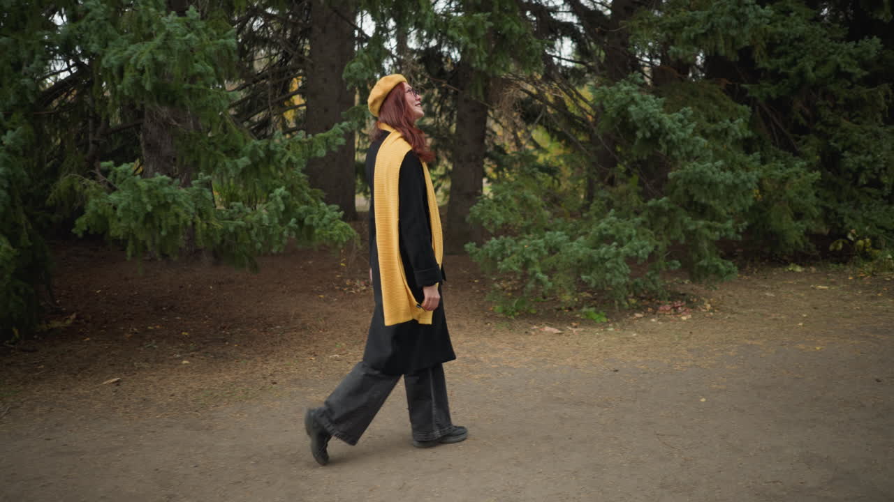 vista lateral de una dama elegante con el cabello rojo largo y ondulado, con un abrigo negro, boina amarilla y bufanda a juego, caminando alegremente a través de un exuberante parque verde adornado con coloridos follajes de otoño