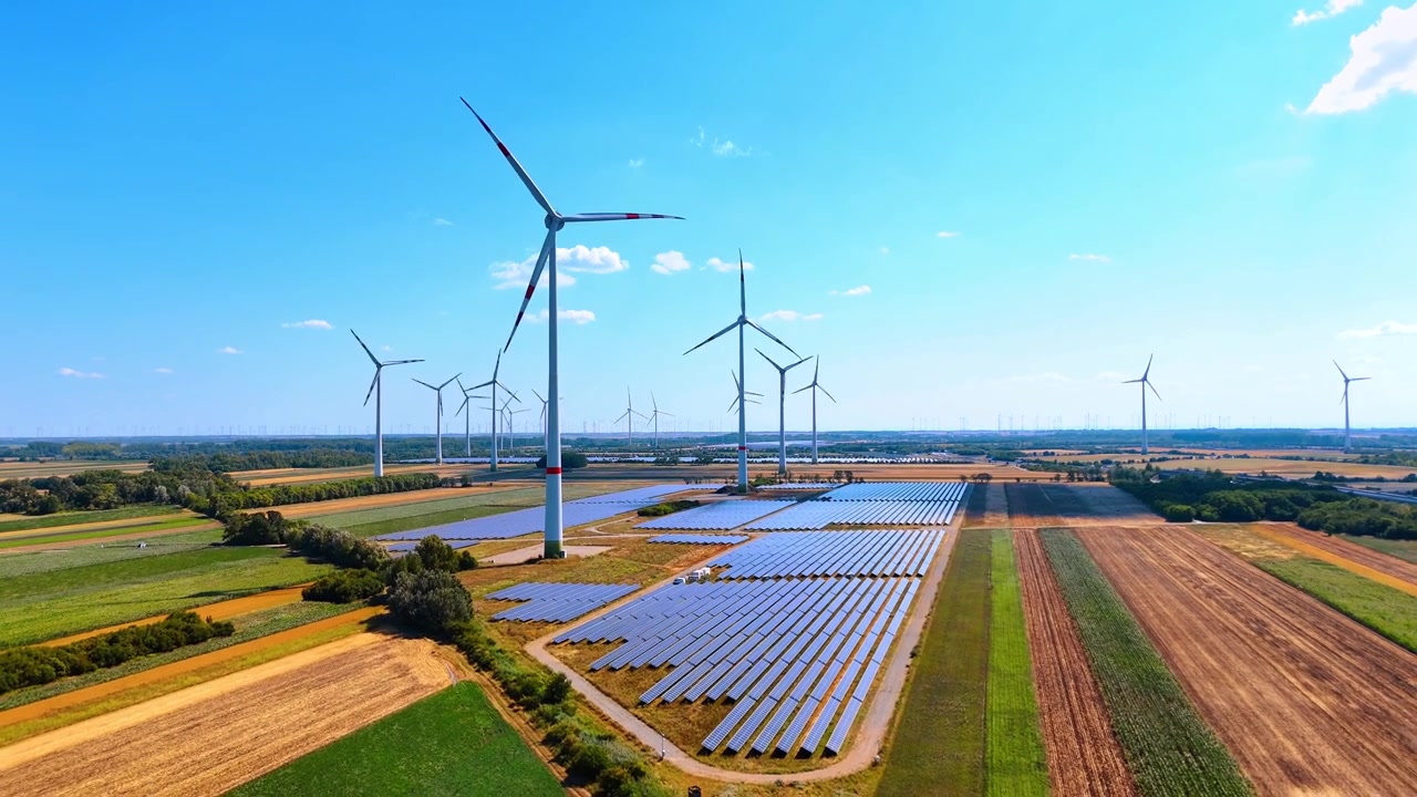 Solcelleanlæg og vindmøller i landskabet. Panoramisk luftfoto af ren energianlæg blandt brune og grønne marker under klar himmel.