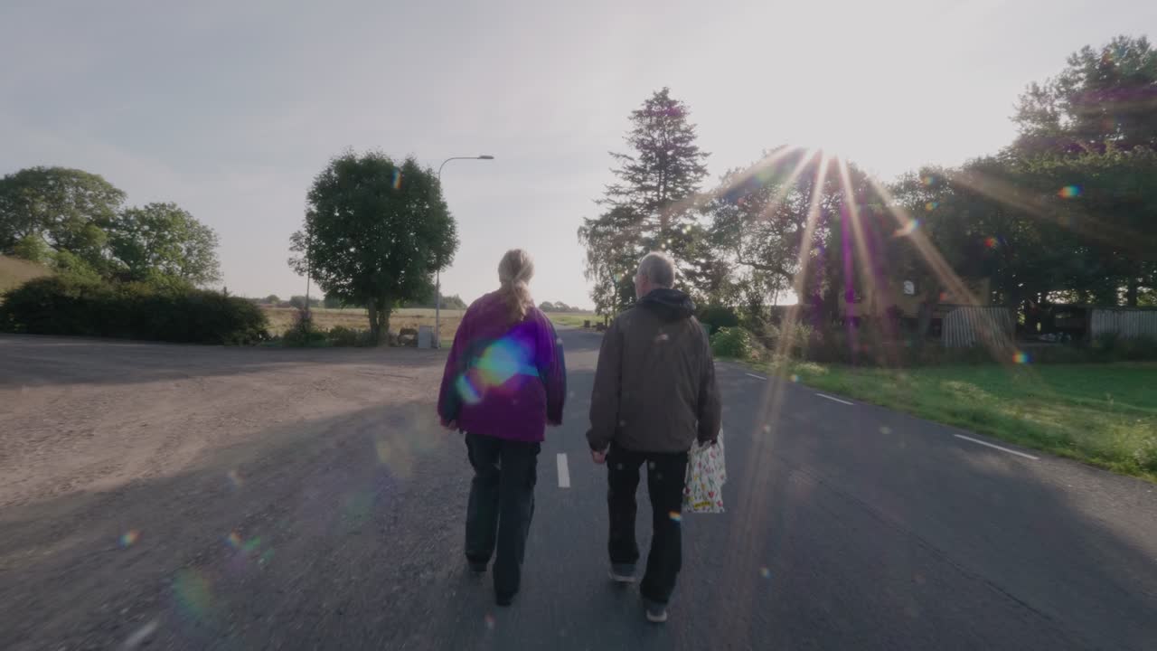 Elderly Couple Walks On Countryside Road With a White Bag In a Small Village, Wide Shot Tracking Forward