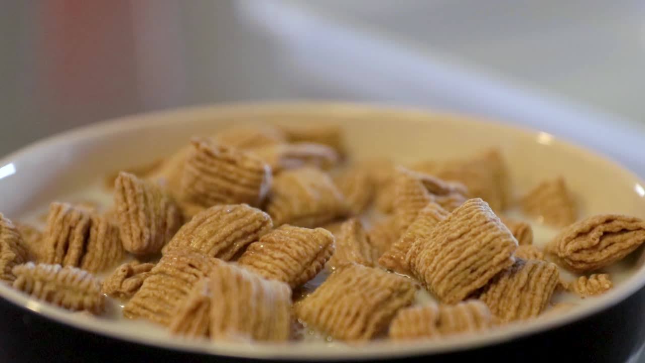 Close up view of milk pouring into a full bowl of tasty cereal while adding a silver spoon.