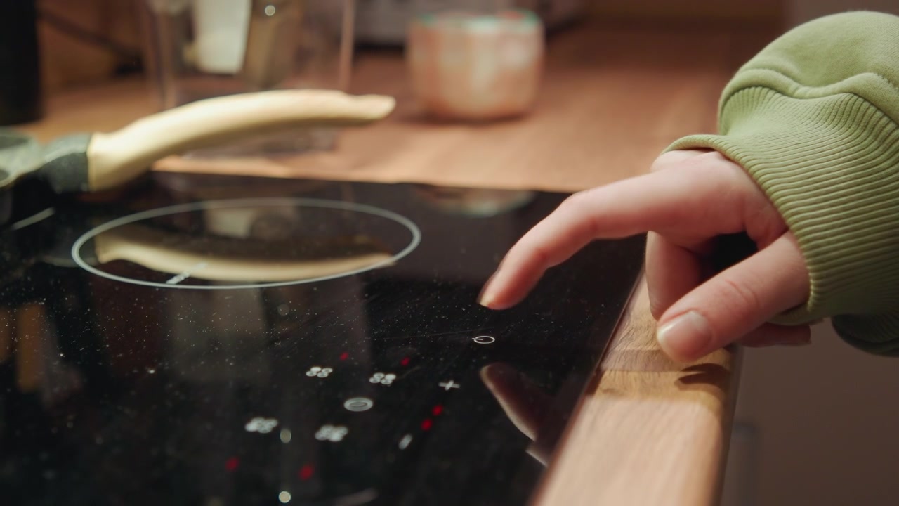 Hand in green sweater scrapes spilled liquid and glass fragments from cracked black ceramic cooktop near wooden countertop, with smears, droplets, reflective surface, and visible damage