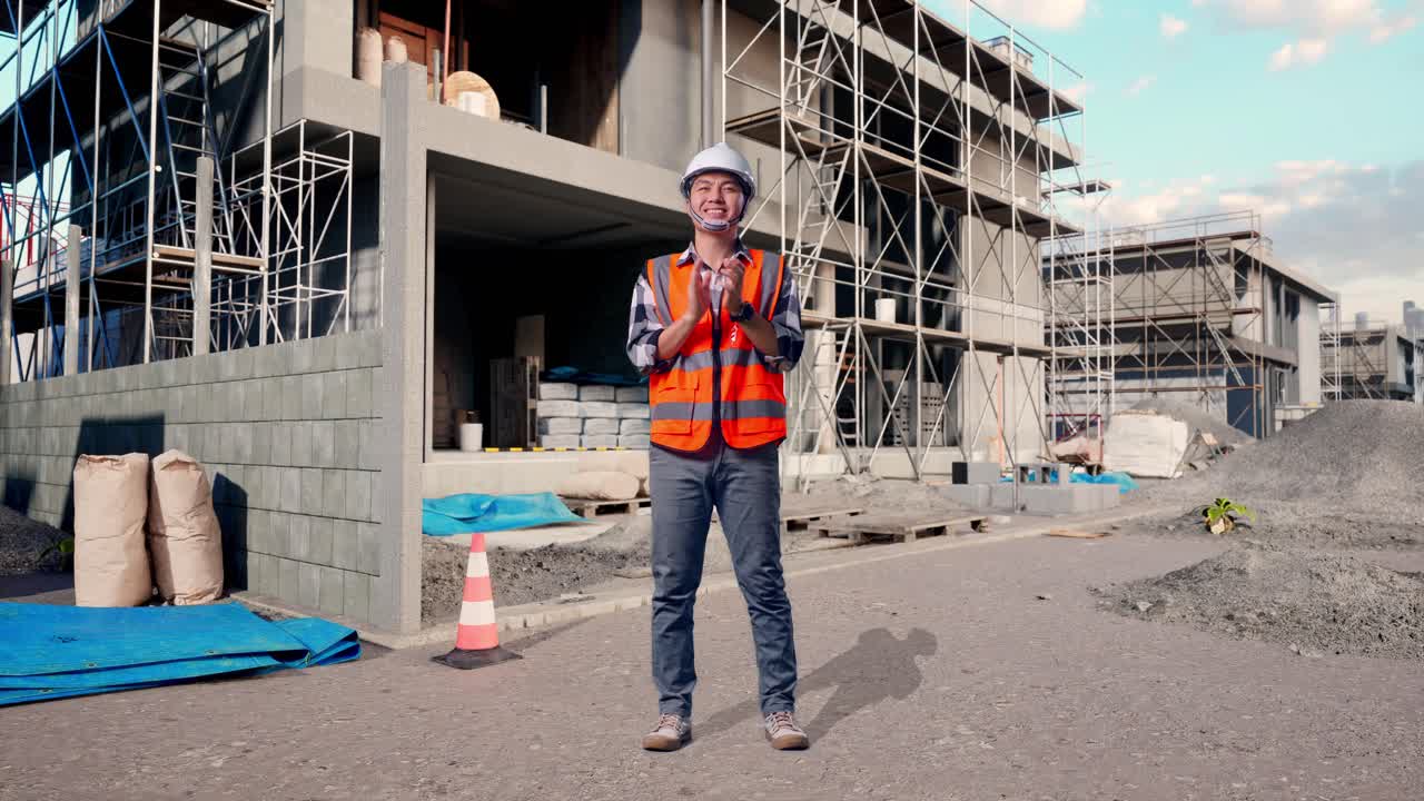 Full Body Of Asian Male Engineer With Safety Helmet Smiling And Clapping His Hands While Standing At Building & Construction Site in Progress to New House