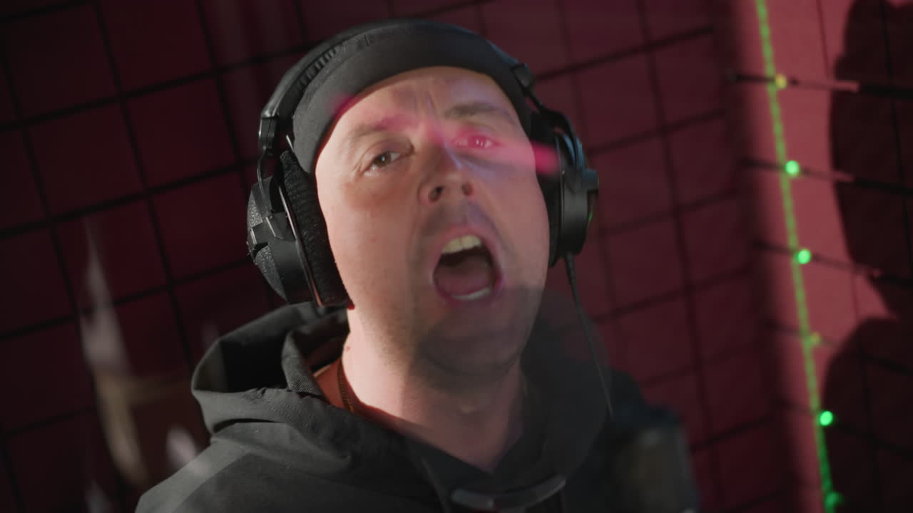 검은 옷을 입은 남자가 음향 방역된 녹음실에서 마이크로 열정적으로 노래를 부르고 있으며, 그의 얼굴은 강렬한 표정을 나타내며 손으로 몸짓을 하며 공연에 완전히 몰입하고 있습니다.