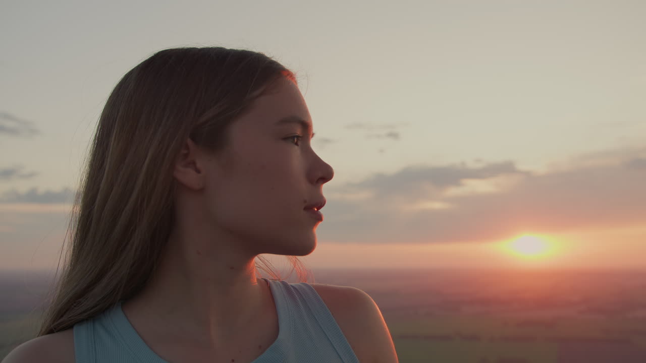 close up profile of young girl turning gaze from distant horizon toward camera while standing in hot air balloon basket under soft sunset sky over expansive fields glowing warm golden light