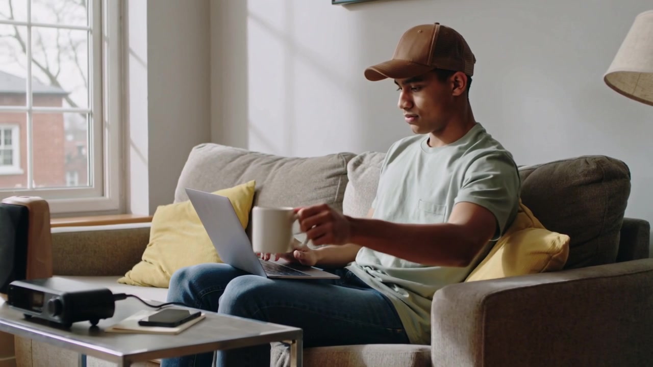 Typing adult wearing green tee and brown cap on sofa at home, grabbing mug using laptop