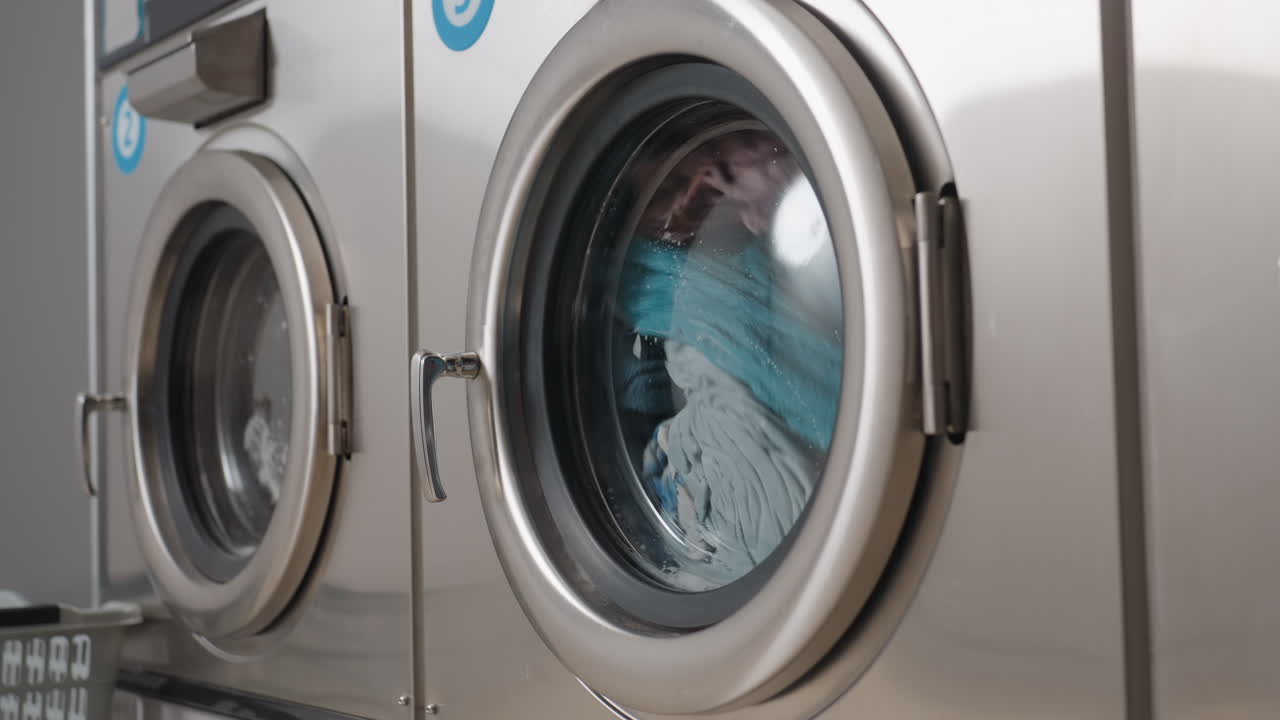 Round view of washing machine components showing front glass door filled with assorted clothes tumbling inside, water and detergent visible through transparent surface during wash cycle in laundromat