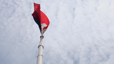 Die-Philippinische-Nationalflagge-Wird-Von-Unten-Neben-Dem-Mast-Gesehen,-Während-Sie-Mit-Etwas-Wind-Und-Watteähnlichen-Weißen-Wolken-Weht,-Während-Die-Kamera-Geneigt-Ist