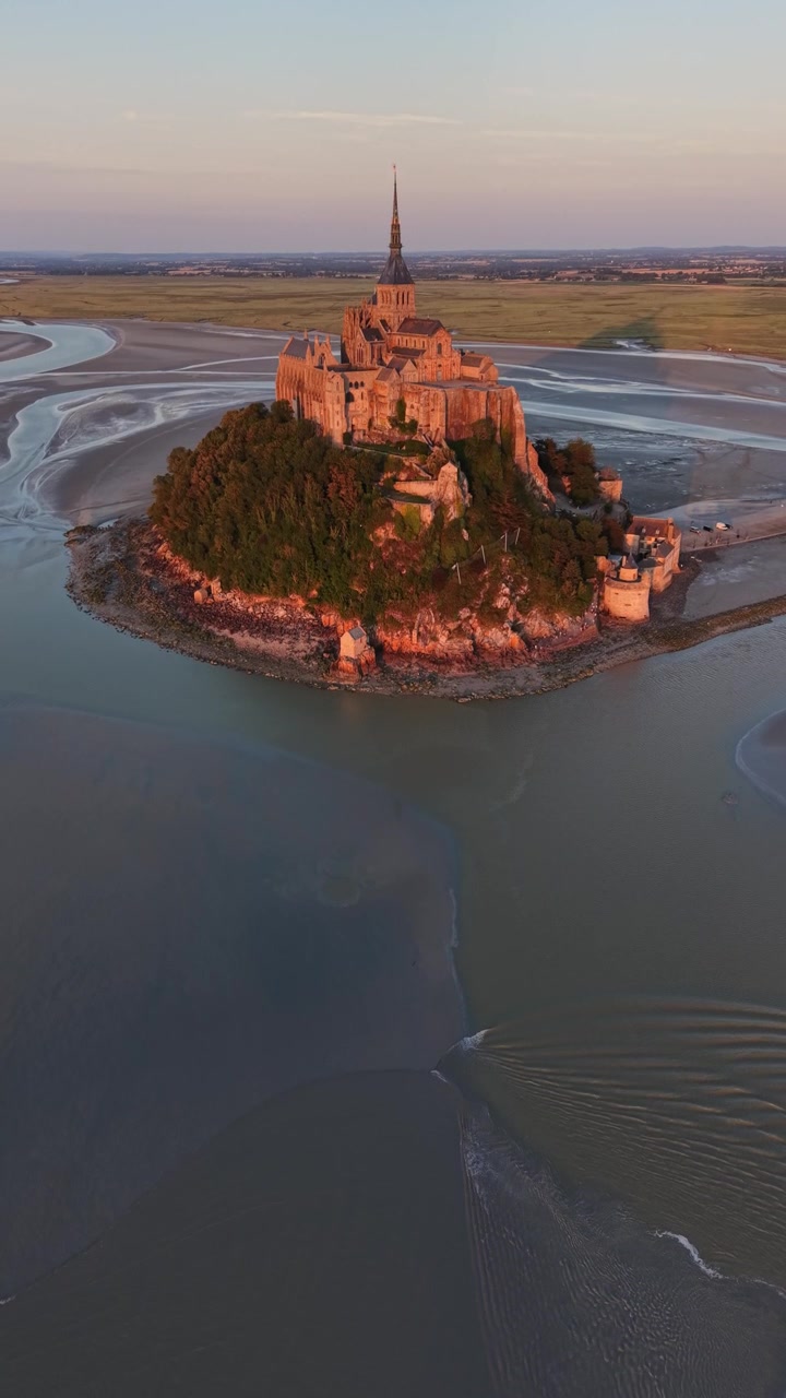 Vertikale droneoptagelser bevæger sig langsomt fremad mod Mont Saint-Michel Kloster under den gyldne time, hvilket fremhæver den middelalderlige ø omgivet af tidevand og kanaler i Normandiet, Frankrig.