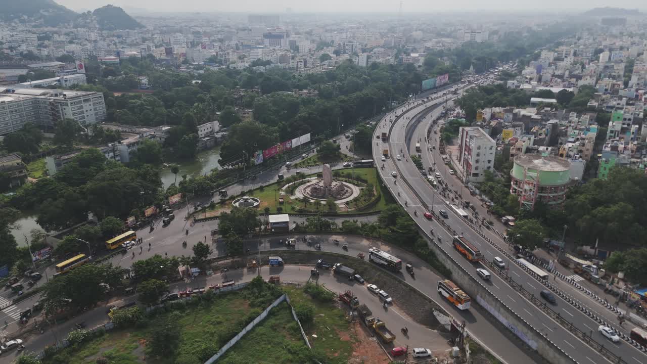In a cinematic aerial spectacle, Vijayawada, a thriving metropolis, unveils its dynamic urban landscape. The majestic Krishna River gracefully the city, while the iconic Dharma Chakra Circle landmark