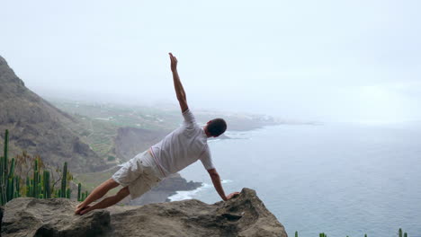 En-Medio-De-Las-Montañas,-Un-Hombre-Practica-Una-Postura-Con-Una-Sola-Mano,-De-Espaldas-A-La-Cámara,-Mientras-Medita-Y-Contempla-El-Océano.