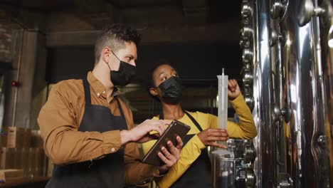 diverse male and female colleague in face masks at gin distillery, checking equipment, using tablet