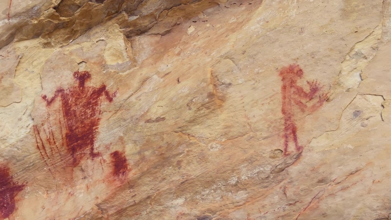 Grande pittogramma antropomorfo rosso dipinto su una parete rocciosa di arenaria, con braccia forti e ondeggianti e figure più piccole sotto.
