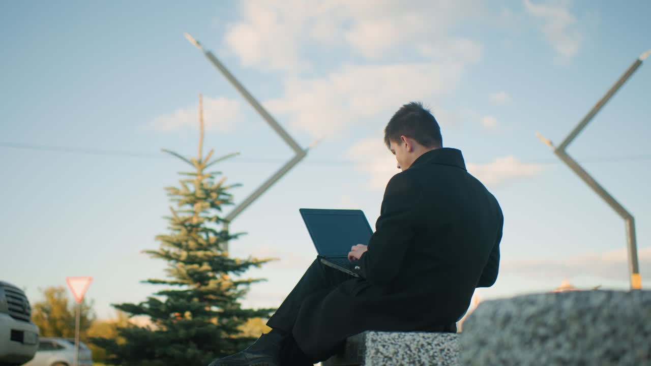 Back view of man seated outdoor operating laptop, with leg crossed, background features parked cars, moving cars, structures and greenery around