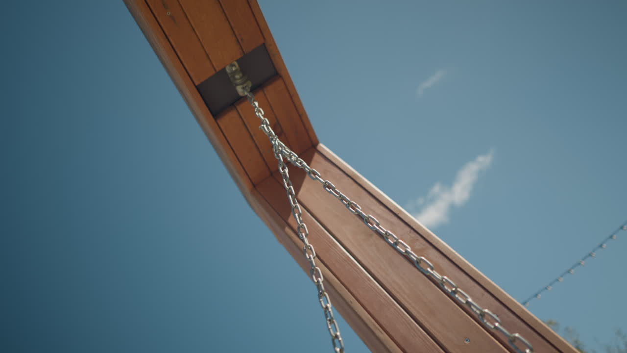 primer plano de columpio y cadena de madera moviéndose suavemente bajo un cielo azul claro y brillante, destacando la simplicidad, la relajación al aire libre y la artesanía en un entorno natural pacífico