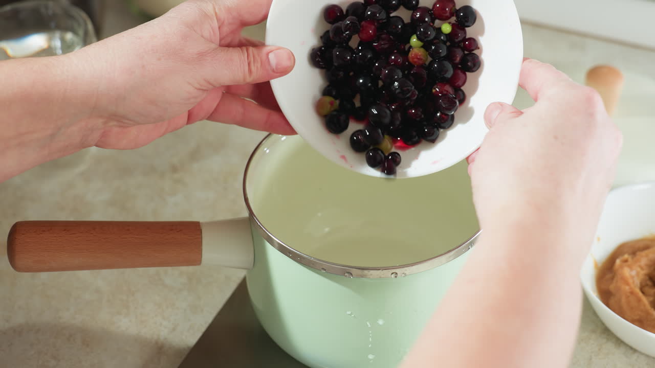 Chef pours black currants into light green pot with wooden handle on digital scale, then sets bowl aside and reaches for second bowl containing mashed ingredients