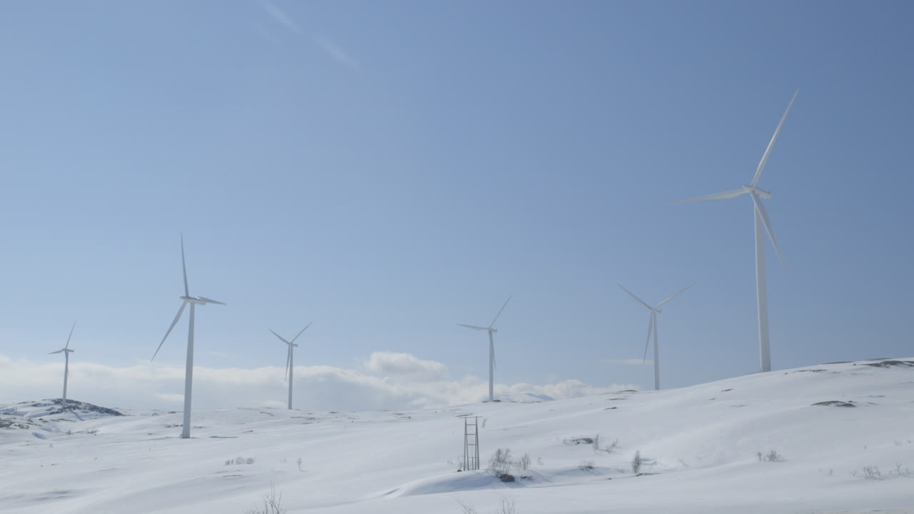 Tripod pan reveals wind turbines farm turning in a bright, snow-covered mountain landscape. Perfect for climate visuals, eco documentaries, or alternative energy editorial projects.