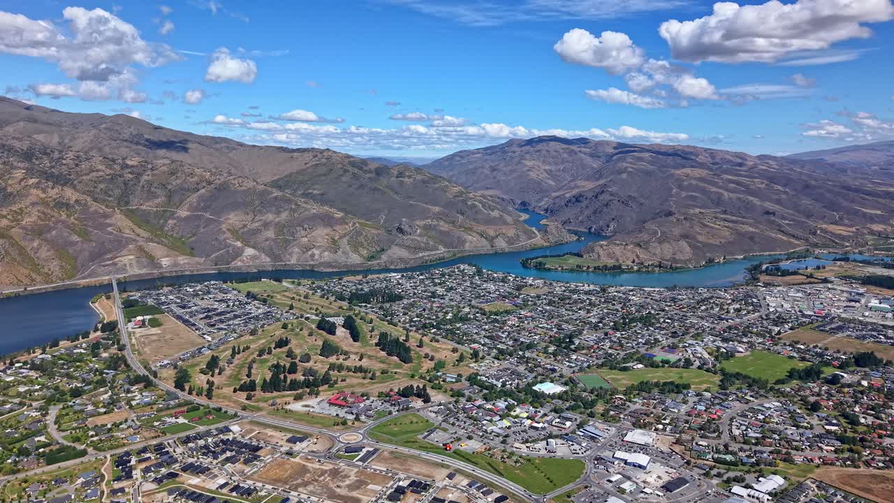 A drone moves forward above Cromwell, showing residential blocks, intersecting roads, the winding Clutha River, surrounding mountains, and a blue sky with scattered clouds