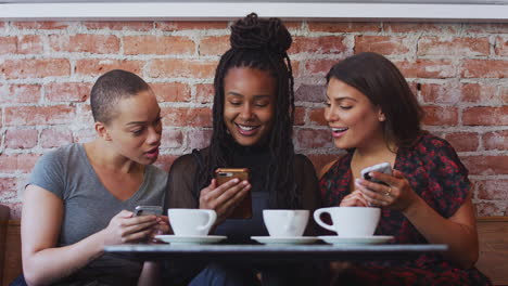 three female friends meeting for coffee sitting at table looking at mobile phones