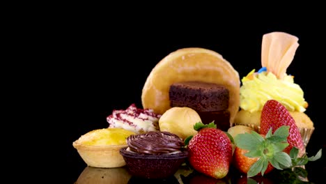 a colorful assortment of donuts, cupcakes, tarts, brownies, and strawberries rotates smoothly under bright studio lighting against a seamless black background, highlighting glossy textures