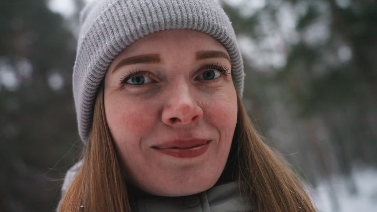Extreme close-up portrait of woman in gray knit beanie and winter coat smiling softly with snowflakes on face and hat, surrounded by blurry snow-covered forest background, capturing peaceful cold weather mood